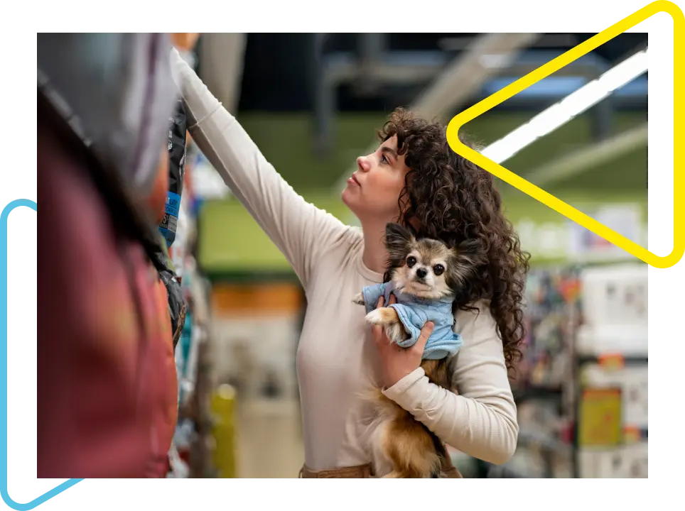 Mulher de cabelo cacheado segurando um cachorro com a mão esquerda enquanto pega um produto no pet shop com a mão direita