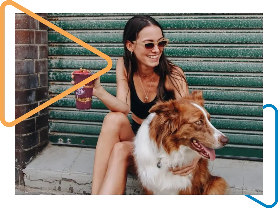 Mulher sentada na calçada de um estabelecimento fechado segurando um cachorro com a mão direita e um copo de açaí Oakberry