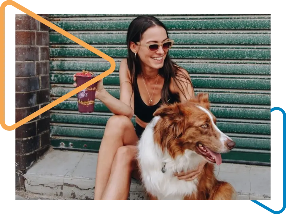 Woman sitting on the sidewalk of a closed establishment holding a dog with her right hand and an Oakberry açaí bowl.