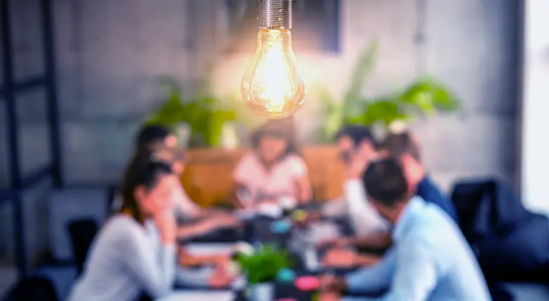 Highlighted light bulb and, in the background, a group of unfocused employees deciding on the creation of an app